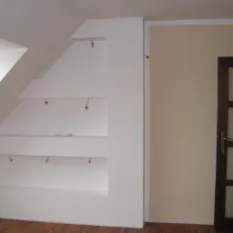 Biała zabudowa wnęk na poddaszu z przygotowanymi kablami do oświetlenia, widoczne okno dachowe i drewniana podłoga.